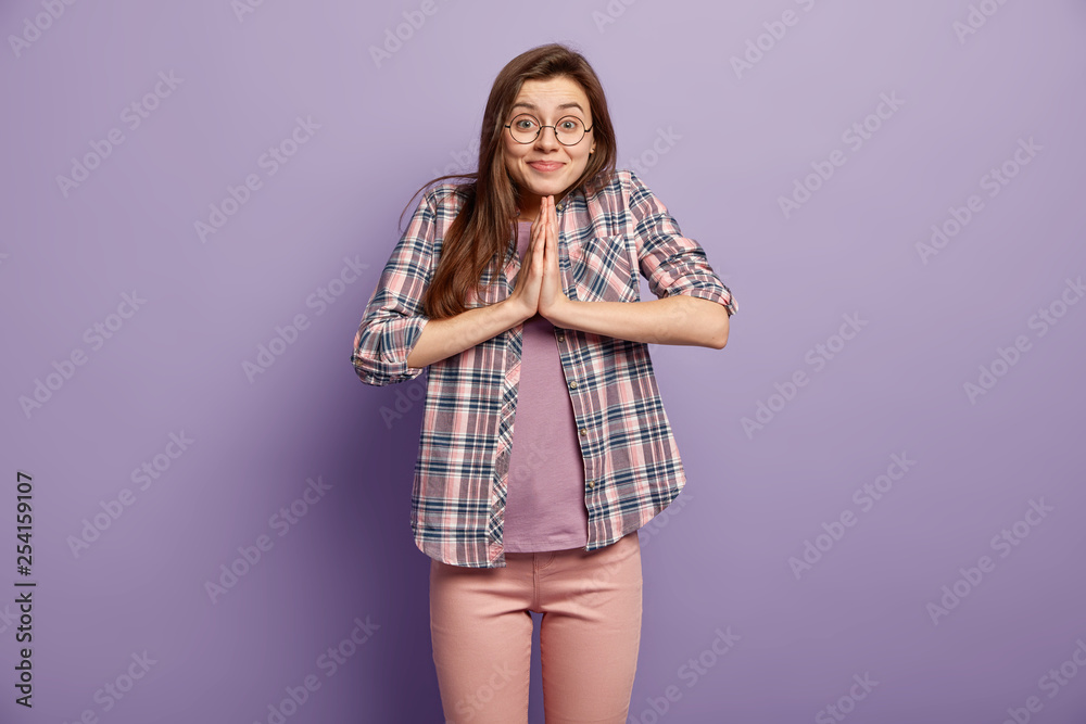 Pleased woman keeps palms pressed together, has gentle smile, asks for favour, gestures indoor, wears checkered shirt and rosy trousers, isolated over purple studio wall, holds hands in pray Pleased woman keeps palms pressed together, has gentle smile, asks for favour, gestures indoor, wears checkered shirt and rosy trousers, isolated over purple studio wall, holds hands in pray