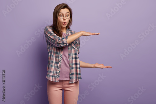 Amazed beautiful young woman surprised which large box she seeks, shows huge object, shapes length with two raised palms, gasps from surprise, demonstrates big thing over purple studio wall.