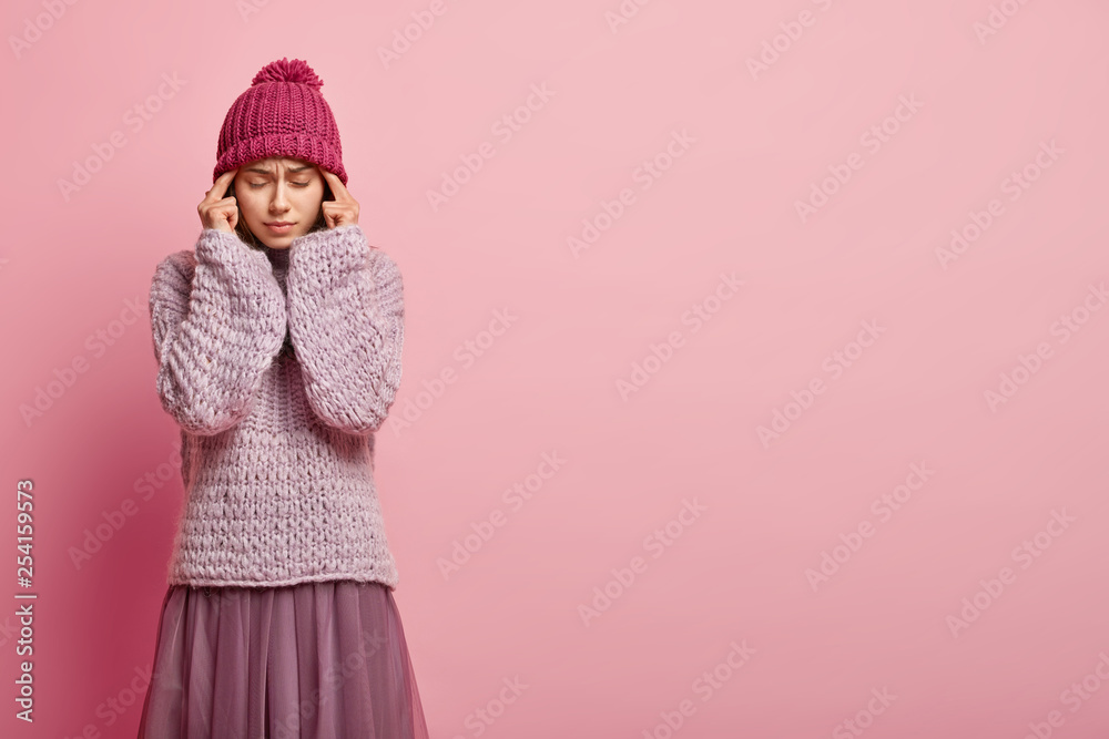 Dissatisfied tired European woman keeps fore fingers on temples, has bad headache after exhausting work, wears winter apparal, closes eyes from pain, isolated over rosy wall with empty space Dissatisfied tired European woman keeps fore fingers on temples, has bad headache after exhausting work, wears winter apparal, closes eyes from pain, isolated over rosy wall with empty space