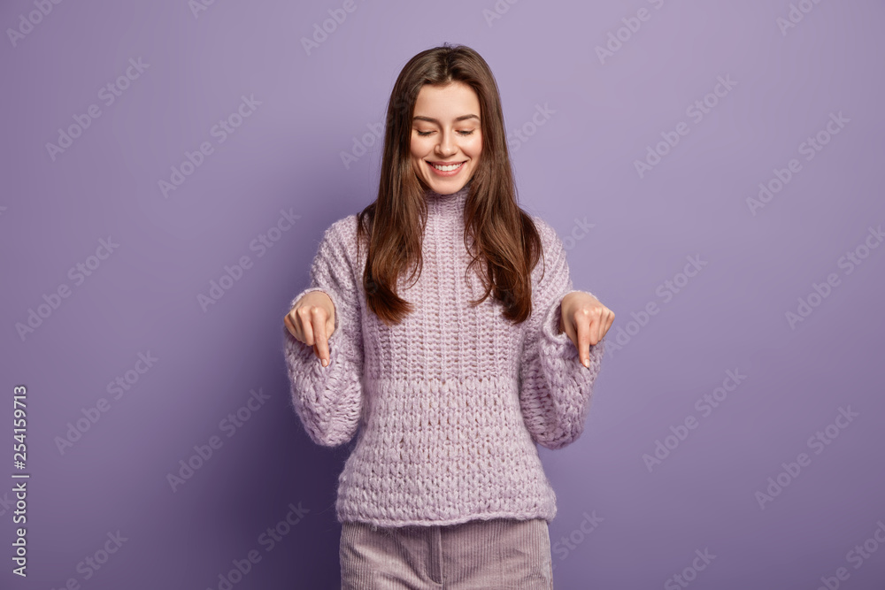 Attractive joyful lovely lady points with both fore fingers down, demonstrates something awesome on floor, dressed in casual knitted sweater, advertises product, wears winter clothing. Monochrome Attractive joyful lovely lady points with both fore fingers down, demonstrates something awesome on floor, dressed in casual knitted sweater, advertises product, wears winter clothing. Monochrome