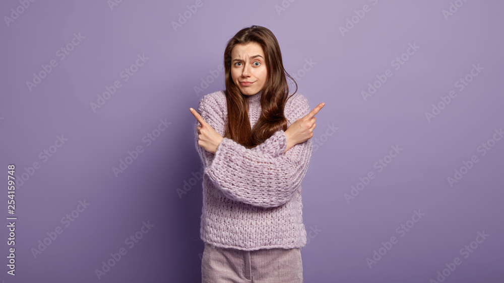 Photo of hesitant woman with puzzled facial expression points sideways, indicates two objects on right and left, cant make choice, faces doubtful situation, has unsure look, wears oversized jumper Photo of hesitant woman with puzzled facial expression points sideways, indicates two objects on right and left, cant make choice, faces doubtful situation, has unsure look, wears oversized jumper