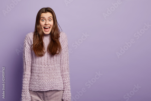 Emotional jovial dark haired woman being on heavens from pleasure, happy to recieve lump sum of money, has cheerful facial expression, wears knitted jumper isolated over purple studio wall, free space