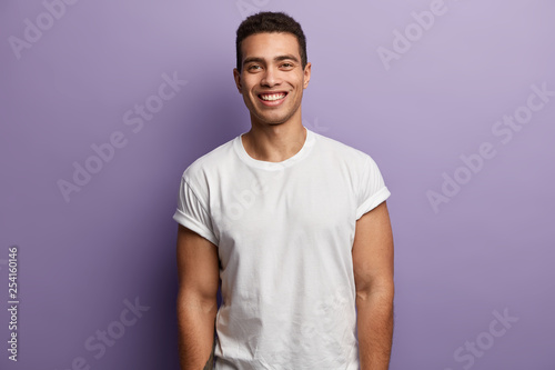 Waist up shot of handsome cheerful man with toothy smile, muscular arms, being in high spirit as spends free time with friends or girlfriend, wears casual white t shirt, isolated over purple wall