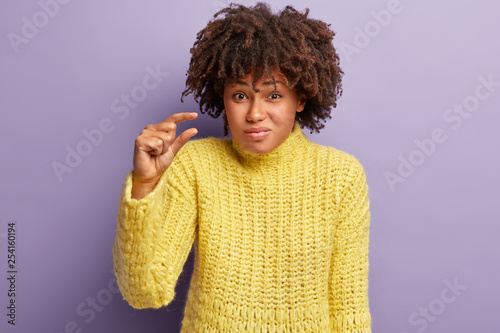 Size matters. Puzzed pretty young woman with Afro hairstyle  has tiny object  demonstrates measure symbol  shapes something small  wears knitted yellow sweater  isolated over purple background
