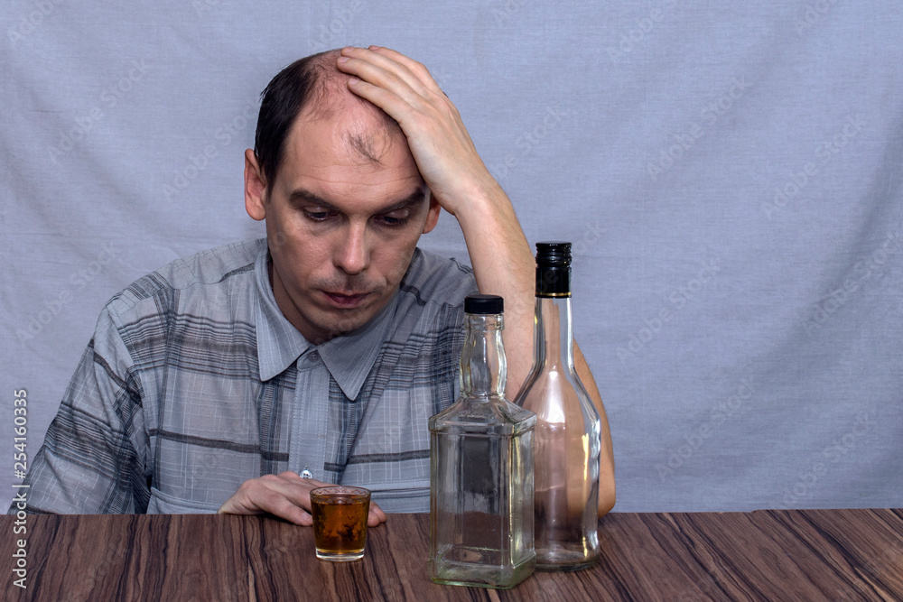 A man sits at the table and looks at a glass with alcohol. Before him stands two empty bottles of alcohol. A man sits at the table and looks at a glass with alcohol. Before him stands two empty bottles of alcohol.