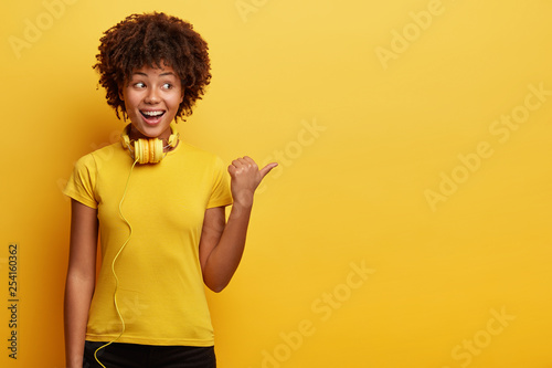 Waist up shot of African American hipster girl being amused by listening music, has modern headphones on neck, points aside, shows way to electronic shop, has cheerful expression, isolated on yellow