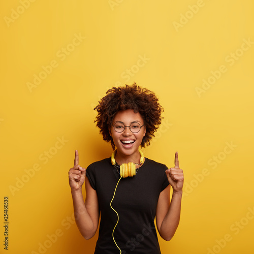 Happy energetic curly young woman points with both fore fingers upwards, being in good mood after party, advertises something above, looks carefree, uses headphones for listening favourite music