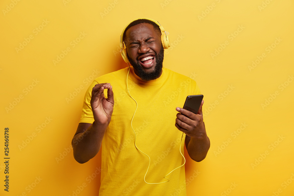 Hipster guy with dark skin, joyful expression, recalls nice memories while listens nice awesome song in headphones, sings loudly, expresses sincere emotions, wears apparel in one tone with background Hipster guy with dark skin, joyful expression, recalls nice memories while listens nice awesome song in headphones, sings loudly, expresses sincere emotions, wears apparel in one tone with background