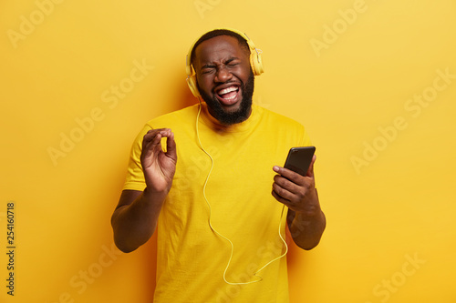 Hipster guy with dark skin, joyful expression, recalls nice memories while listens nice awesome song in headphones, sings loudly, expresses sincere emotions, wears apparel in one tone with background