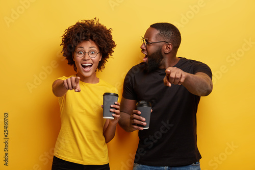 Happy dark skinned woman and her boyfriend being emotional, surprised, point at camera, laugh at something funny, drink takeaway coffee, enjoy free time together, react on something astonishing