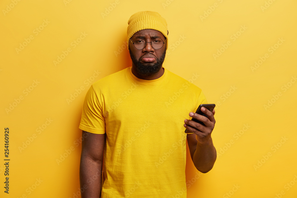 Dissatisfied dark skinned plump guy wastes time with modern technologies, holds cell phone, upset not to recieve message from important person, wears yellow hat and t shirt, feels dejected. Monochrome Dissatisfied dark skinned plump guy wastes time with modern technologies, holds cell phone, upset not to recieve message from important person, wears yellow hat and t shirt, feels dejected. Monochrome