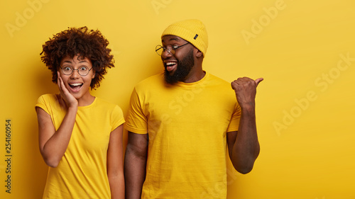 Fotografi Studio shot of cheerful woman hears advice from boyfriend who tells something and points aside with thumb, dressed in similar yellow t shirts, being friendly team of coworkes