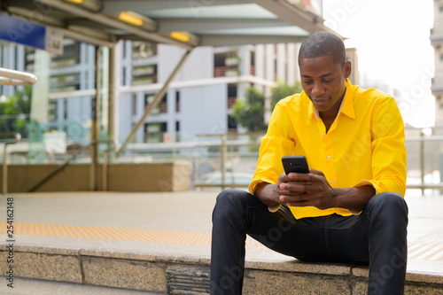 Young bald African businessman using phone while sitting outside the subway train station