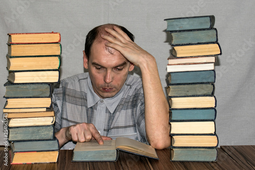 A man is reading a book holding his head with one hand. On the table are two large stacks of books.