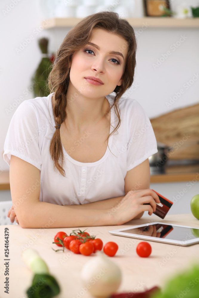 Young woman looking for a new recipe for cooking in a kitchen. Housewife is making online shopping by tablet computer and credit card Young woman looking for a new recipe for cooking in a kitchen. Housewife is making online shopping by tablet computer and credit card