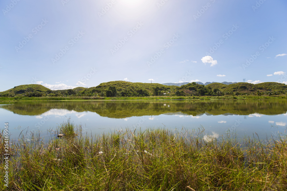 Fishpond nature scenery in sunlight with sunbeams and reflections of blue sky, white clouds and green trees in the water