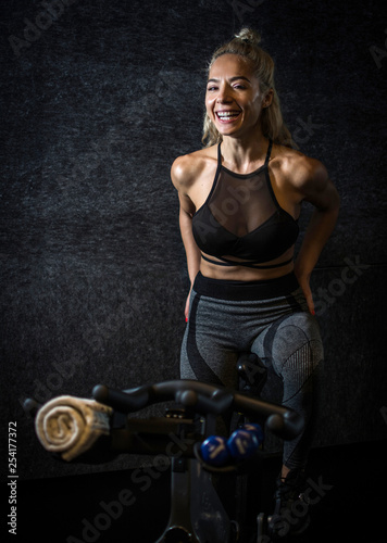 Cheerful sportswoman on exercise bike against dark background