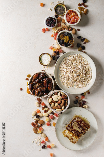 Variety of dried fruits, nuts, honey and oat flakes in ceramic bowls for cooking homemade healthy breakfast muesli or granola energy bars over white texture background. Flat lay, space.