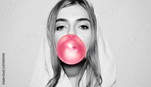 Portrait of young playful blonde girl dressed in hoodie standing isolated over pink background while blowing bubble with chewing gum. Woman looking at camera photo
