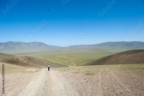 A boy looks at a steppe road through a valley in Mongolia, an eagle flies above him in the sky