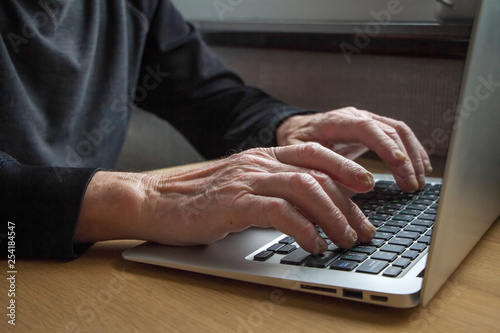 The hands of an elderly person working on a laptop keyboard, typing, close up photo.