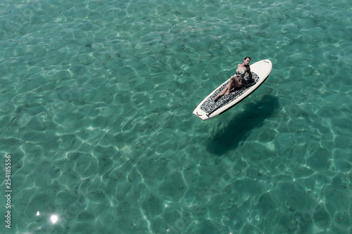 A beautiful young woman relaxes on a SUP board in the sea near the island. Drone shot