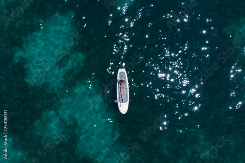 A beautiful young woman relaxes on a SUP board in the sea near the island. Drone shot