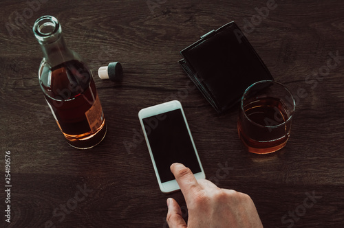 A man presses a finger on a mobile phone. Next on the table is a glass of whiskey, a bottle of whiskey and a purse.