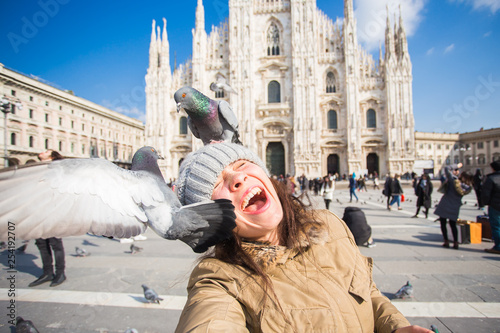 Obraz na płótnie Winter travel, vacations and birds concept - Young happy woman tourist with funny pigeons making selfie photo in front of the famous Duomo cathedral in Milan