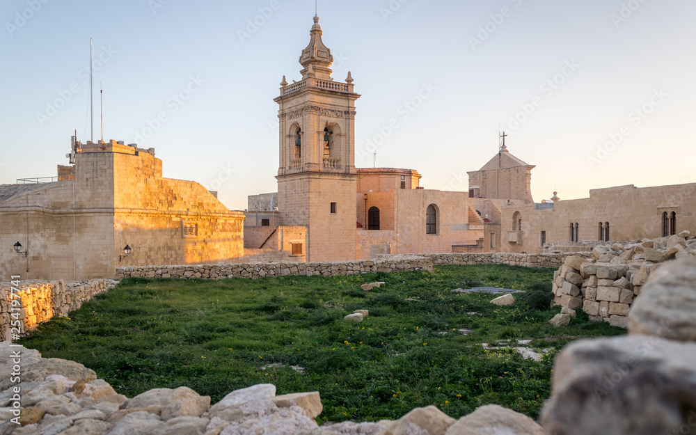 Detail view on the old, historical St. Joseph's Chapel inside the Citadel of Victoria inside antique ruins walls with grass field and sandstones in the foreground on Gozo, Malta Detail view on the old, historical St. Joseph's Chapel inside the Citadel of Victoria inside antique ruins walls with grass field and sandstones in the foreground on Gozo, Malta
