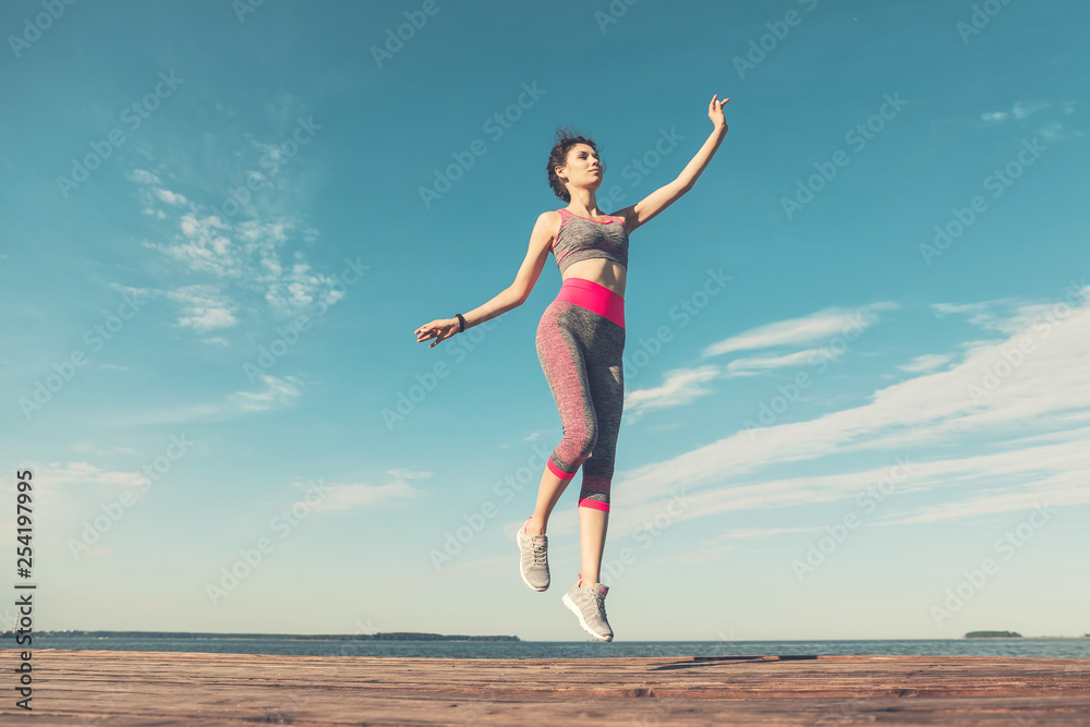 Fototapeta premium Sporty young girl in the morning on pier in jump, practicing yoga. Woman do gymnastics outdoors. Health and Yoga Concept. Female lifestyle meditation for fitness lifestyle the nature background