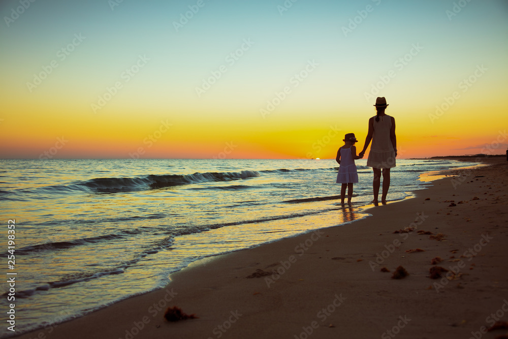 mother and daughter tourists on ocean coast at sunset walking