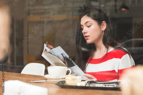Cute brunette girl sits in a cafe with a cup of coffee and a dessert on the table, holds a magazine and reads in her hands. Portrait of a girl resting in a cafe.