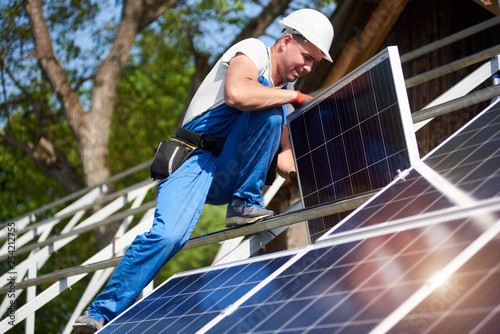 Professional technician adjusting heavy solar photo voltaic panel to high metal platform on blue sky and green tree background. Stand-alone solar system installation, green energy production concept.