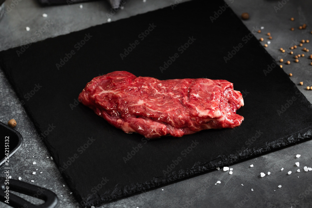 Raw meat, beef butcher or machete steak on black Slate board with Grill pan, knife with spices on a gray table