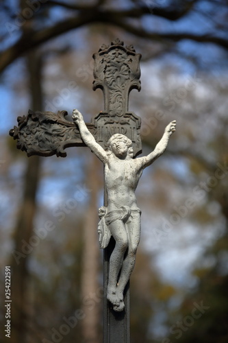 Old made of metal Christian cross with broken one arm, with Jesus Christ figure  on it, in background trees in soft focus