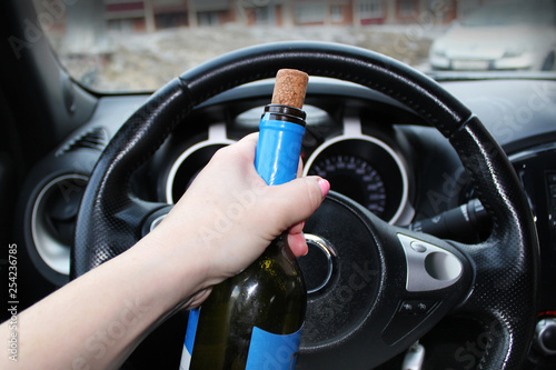 Hand with a bottle of alcohol near the car steering wheel. Drinking and driving. © Maria