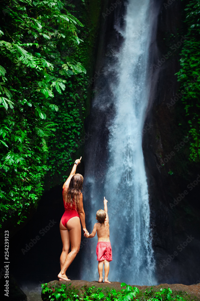 In tropical jungle happy mother with kid stand under waterfall, explore rainforest nature. Travel adventure, hiking activity with child. Lifestyle on family summer vacation and weekend walking tour.