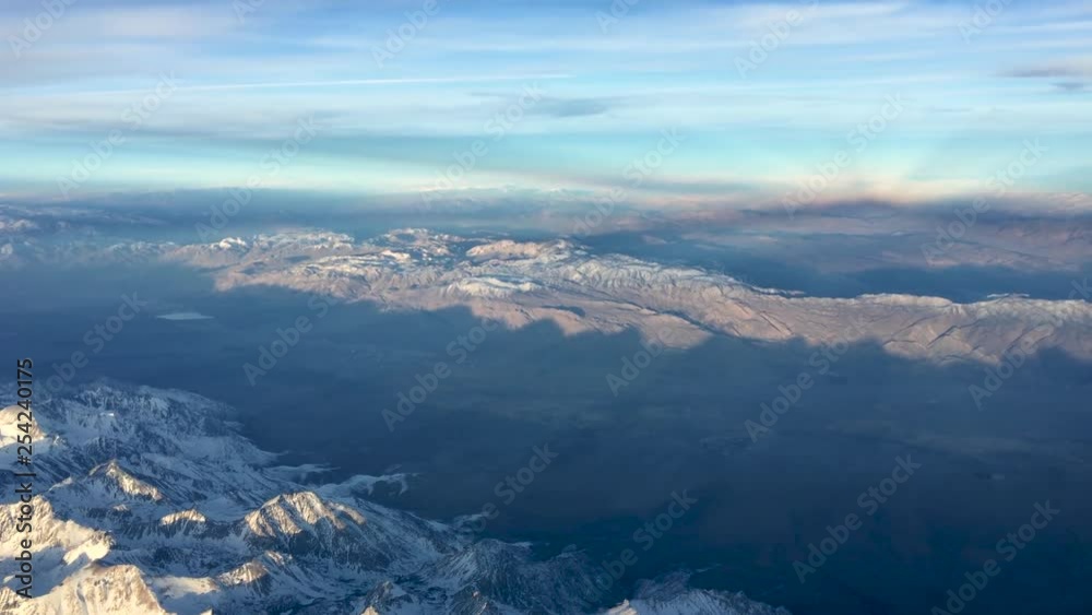 Clean aerial view of snow capped mountains above valley from plane at altitude looking out left side,