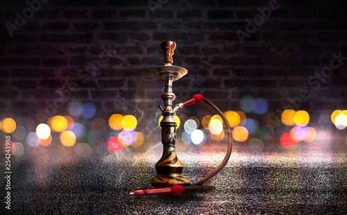 Hookah with smoke on the background of an empty brick wall, wet asphalt and colored neon light, blurry lights of the night city, bokeh