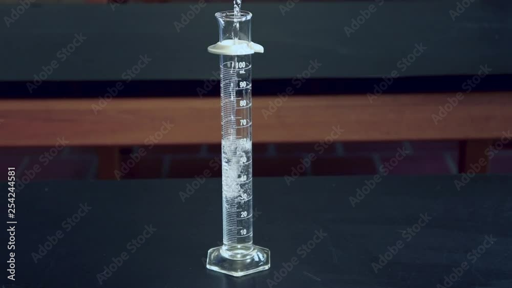 Clear liquid being poured into a 100mL graduated cylinder on a black