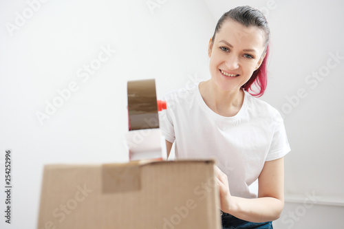 Young beautiful brunette girl in a white t-shirt packs cardboard boxes with a dispenser and adhesive scotch adhesive tape. The concept of moving to a new home.