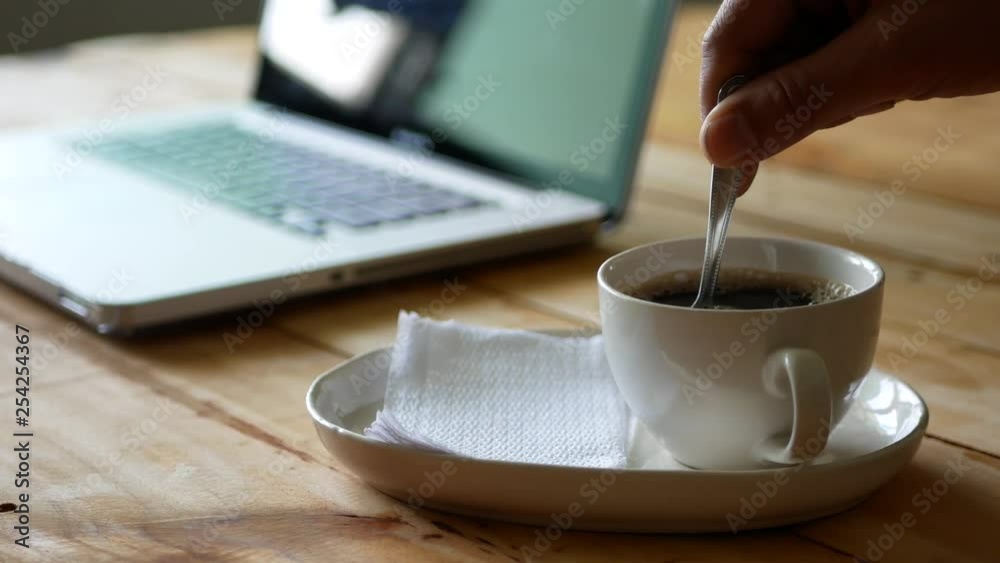 Hot black coffee in a coffee cup being stirred with a computer notebook in the background