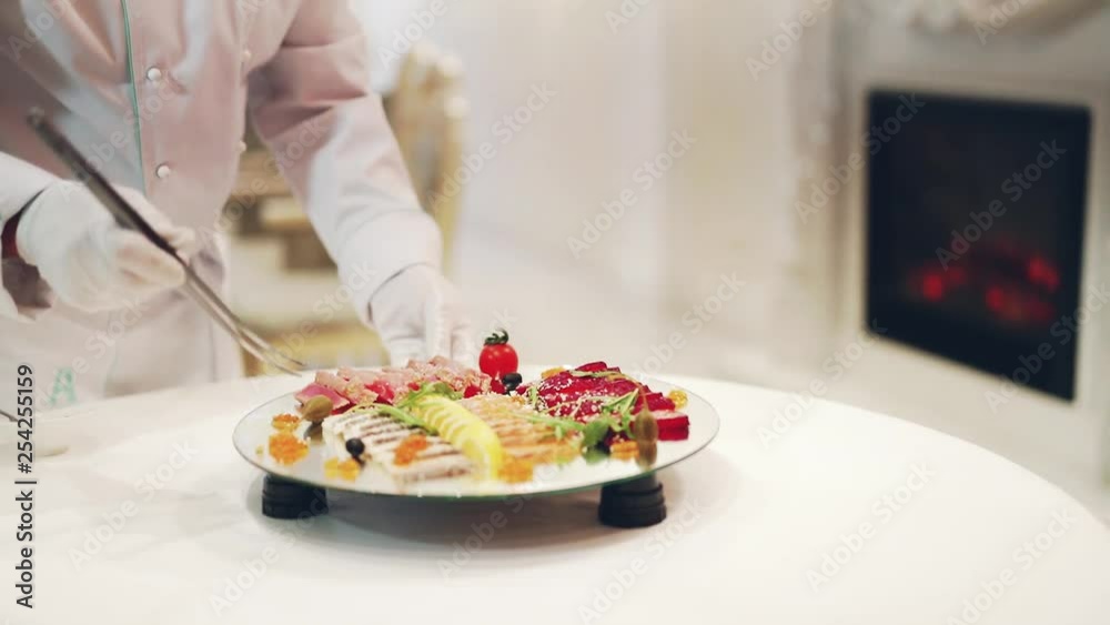 Unrecognizable male cook in white gloves decorates plate with delicious food on a glass turntable. Professional cook puts slicing products prettily for guests. Unrecognizable male cook in white gloves decorates plate with delicious food on a glass turntable. Professional cook puts slicing products prettily for guests.