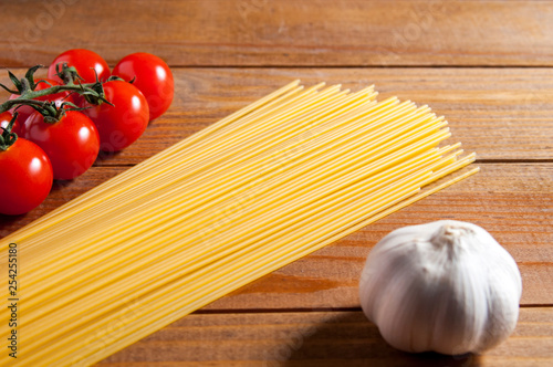 Spaghetti, cherry tomatoes and bulb of garlic on a brown wooden table. Tomatoes to the left and garlic to the right of spaghetti. Close-up.
