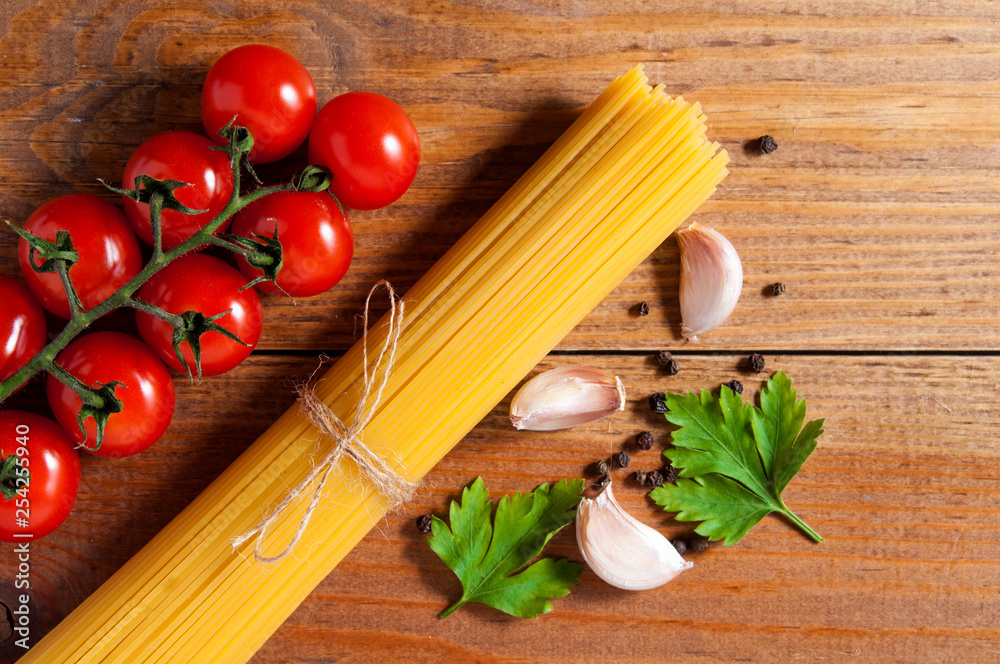 Bunch of raw spaghetti tied with rope, tomatoes cherry, slices of garlic, parsley leaves and pepper on brown wooden background. Top view. Bunch of raw spaghetti tied with rope, tomatoes cherry, slices of garlic, parsley leaves and pepper on brown wooden background. Top view.