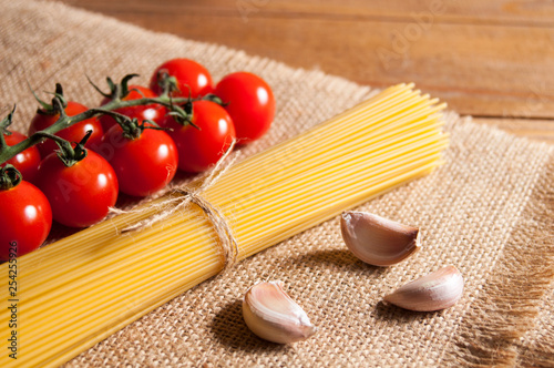 Bunch of raw spaghetti tied with rope, tomatoes cherry and slices of garlic on burlap, all on brown wooden background.