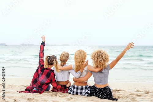 Four multicultural teenage girls hugging and looking at ocean while sitting on the beach. Backs turned.