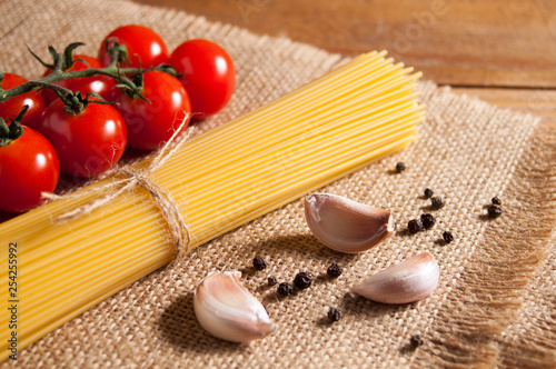 Bunch of raw spaghetti tied with rope, tomatoes cherry, pepper and slices of garlic on burlap, all on brown wooden background.