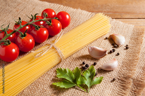 Bunch of raw spaghetti tied with rope, tomatoes cherry, slices of garlic, parsley leaves and pepper on burlap, all on brown wooden background.